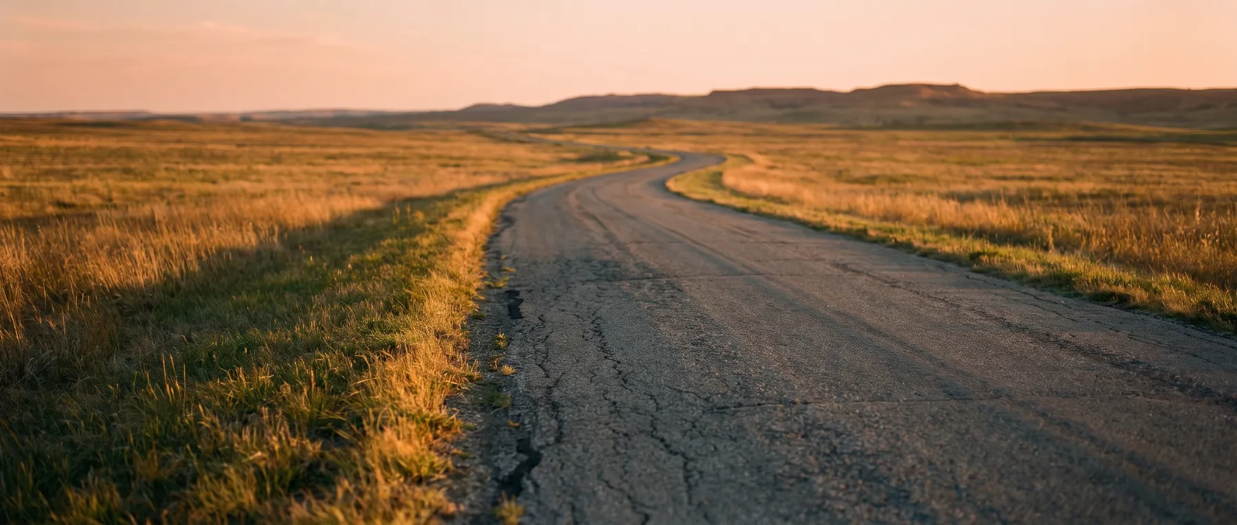 A single-lane road curving through open grassland toward low hills at golden hour.